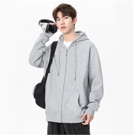 Front view of a man showcasing a casual light grey zip-up hooded jacket with soft drawstrings, front slash pockets, and ribbed cuffs paired with dark trousers.